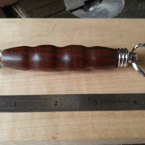 May include: A handcrafted bottle opener with a dark brown, ridged wooden handle and a silver metal ring. The opener features a silver metal triangle design on one end and a metal ring on the other. A metal ruler is next to the bottle opener.