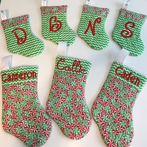 May include: Seven personalized Christmas stockings with green and white peppermint candy designs. The stockings are embroidered with the names Cameron, Callie, Gaden, B, N, D, and S.