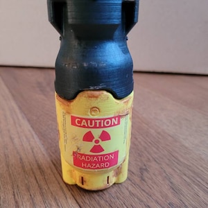 May include: A small yellow container with a black top and a red radiation symbol. The container has the words "CAUTION" and "RADIATION HAZARD" printed on it.