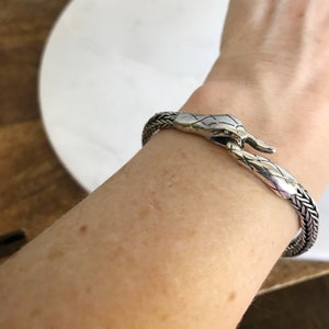 May include: A silver snake-shaped bracelet with a woven design. The bracelet is open and has a clasp that is shaped like a snake's head.