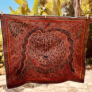 May include: A square textile wall hanging featuring a central embroidered leopard face surrounded by a dark snake, leafy branches, and geometric floral patterns in shades of rust, brown, black, and deep red, suspended by wooden clothespins.