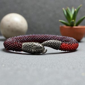 Könnte beinhalten: Ein Perlenarmband mit einem Farbverlauf von Dunkelrot zu Hellrot. Die Enden des Armbands sind mit metallisch silberfarbenen Schlangenkopfverschlüssen versehen, die ineinandergreifen und eine durchgehende, kreisförmige Form mit Textur bilden.