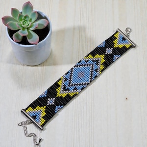 May include: A beaded bracelet featuring a geometric design in blue, yellow, and white against a black backdrop. It has a silver clasp and chain. A small potted succulent plant is visible in the background.