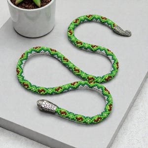 May include: A long, flexible snake-shaped necklace crafted from small green, white, and brown beads arranged in a zigzag pattern. The piece features a detailed silver-toned metal head at one end and a matching silver-toned tail at the other end.