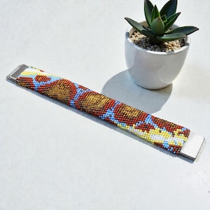 Peut inclure: Un bracelet de perles avec un motif coloré de perles rouges, dorées, bleues et jaunes. Le bracelet a des fermoirs en métal argenté. Un petit pot blanc avec une plante succulente est en arrière-plan.