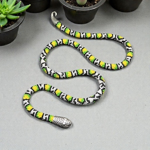 May include: A flexible snake-shaped accessory crafted from small beads in a repeating pattern of yellow, lime green, black, and white. The piece features a metallic silver-toned head and a matching silver-toned tail, both with textured scale details.
