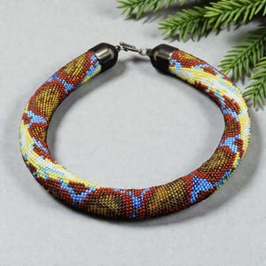 Op de afbeelding: Een kralenketting met een buisvormig ontwerp. De ketting heeft een patroon van rode, gele, blauwe en witte kralen. De uiteinden van de ketting hebben zwarte metalen doppen en een zilveren sluiting. De ketting is ongeveer 40 cm lang.