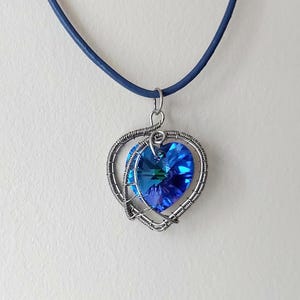 Könnte beinhalten: Herzförmiger Anhänger mit einem leuchtend blauen Kristall in der Mitte, eingefasst in einem silbernen Drahtrahmen. Der Anhänger hängt an einem dunkelblauen Band. Der Kristall reflektiert das Licht und erzeugt einen funkelnden Effekt. Schmuck auf weißem Hintergrund.