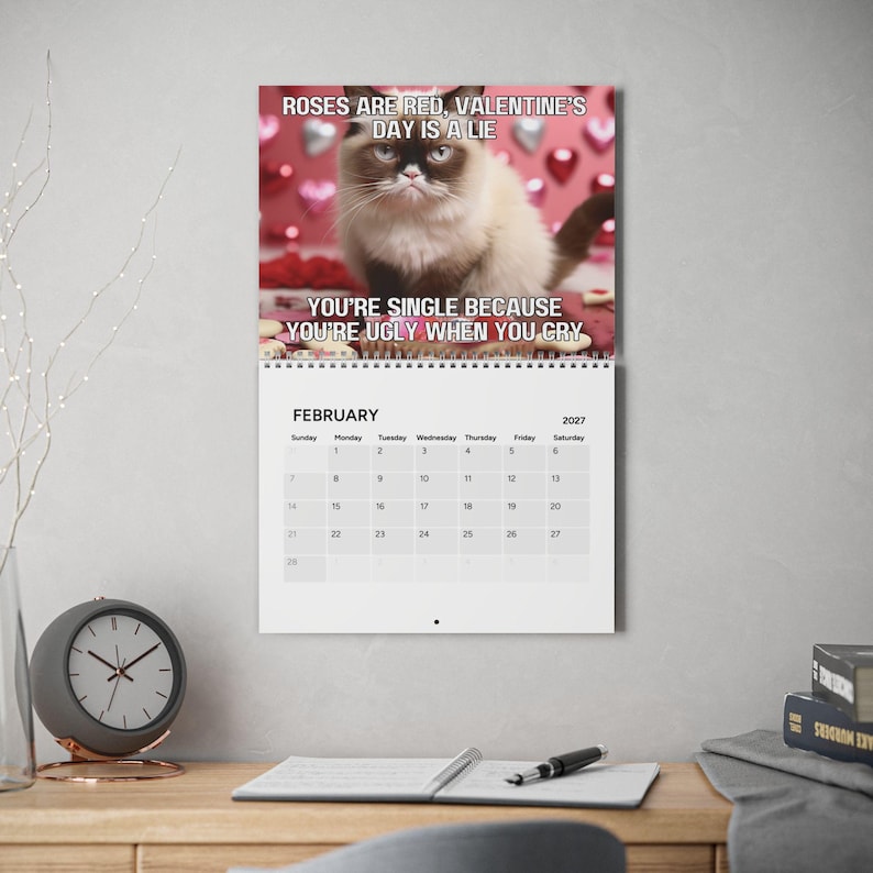 May include: A wall calendar for February 2027 featuring a photograph of a grumpy cat against a red background with heart shapes. The text reads: ROSES ARE RED, VALENTINE'S DAY IS A LIE. YOU'RE SINGLE BECAUSE YOU'RE UGLY WHEN YOU CRY.