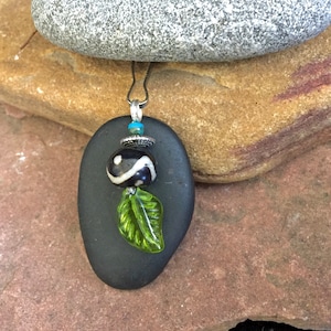 May include: A black stone pendant with a green glass leaf charm and a black and white patterned bead. The pendant is hanging from a silver chain.