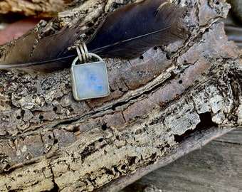 May include: A square pendant with a blue and white moonstone set in silver. The pendant is resting on a piece of driftwood with a black feather.