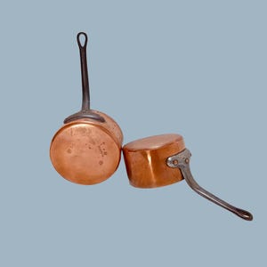 Puede incluir: Dos cacerolas de cobre con asas de metal oscuro. Una tiene un asa larga y base redonda, la otra un asa más corta y forma rectangular. El cobre tiene un cálido tono marrón rojizo.