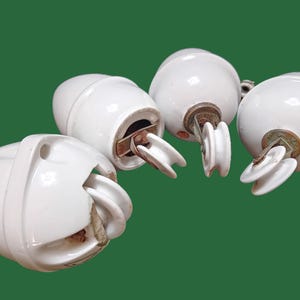 May include: Four white ceramic insulators with metal hardware, arranged against a green background. The insulators have a rounded shape with a groove for wire placement. The metal components show signs of age.