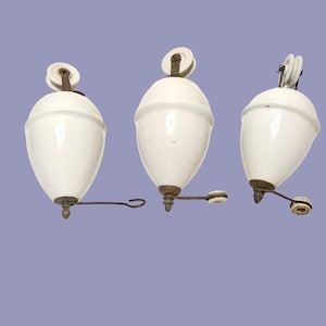 May include: Three white ceramic counterweights with a smooth, glossy finish. Each weight features a metal pulley at the top and a metal arm extending from the bottom, ending in a small white ceramic ring or hook component for attachment.