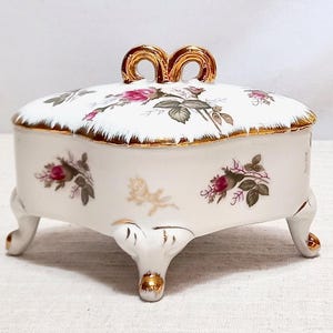 May include: A white porcelain trinket box with a heart-shaped lid featuring a gold-trimmed handle. The exterior is decorated with pink rosebud motifs and green leaves, accented by gold detailing on the scalloped edges and three ornate feet.