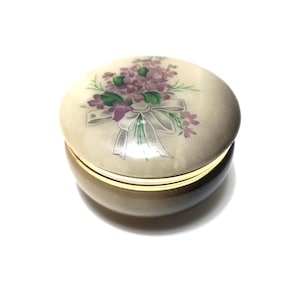 Puede incluir: Una pequeña caja redonda para baratijas con tapa. La tapa presenta un diseño floral con flores moradas y una cinta blanca. La caja tiene una banda dorada alrededor del centro y una base color crema.