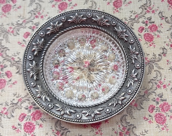 May include: A circular decorative dish featuring a clear, pressed glass center with a starburst and dot pattern. The glass is encased in a dark, antiqued silver-toned metal rim decorated with a repeating embossed floral and leaf motif.