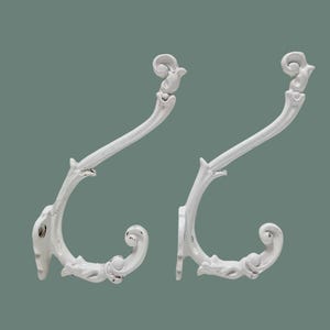 Puede incluir: Dos perchas de pared decorativas blancas con un diseño vintage ornamentado. Los ganchos tienen una forma curvada con detalles de volutas y un acabado envejecido, perfectos para añadir un toque de elegancia a cualquier espacio.
