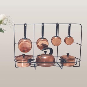 May include: A set of copper cookware hanging on a black metal rack. The set includes a kettle, two small pots, and four saucepans.