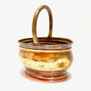 Może przedstawiać: Mały, owalny kwietnik z mosiądzu z miedzianym dnem i pojedynczym uchwytem. Kwietnik ma fakturę młotkowaną i zużyty, antyczny wygląd.