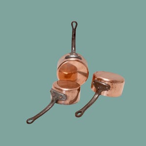 Pode incluir: Um conjunto de três panelas de cobre com superfícies metálicas polidas e cabos de ferro escuro rebitados. Cada panela apresenta um corpo cilíndrico com base plana e um cabo longo e fino que se estende a partir da lateral da peça.