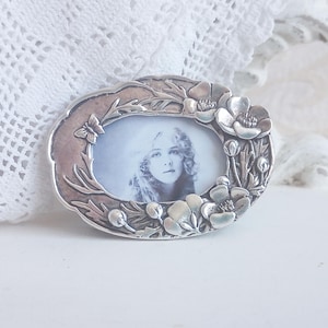 May include: An oval silver-toned metal picture frame featuring a raised relief design of blooming flowers, leaves, and a small butterfly. The frame contains a black and white portrait of a person with wavy hair, set within a decorative border.