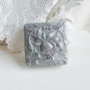 May include: A rectangular, silver-colored metal box with a floral design. The box features a detailed relief of a fuchsia flower surrounded by leaves and vines. The design is intricate and ornate, with a vintage aesthetic.