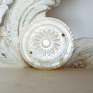 May include: A round, mother-of-pearl button with a floral design. The button features a central flower with surrounding petal details and a rope-like border. Two small holes are visible near the edge. The button is set against a white, textured background.