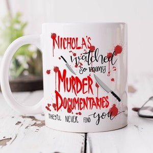 Op de afbeelding: Witte keramische koffiemok met een rode afbeelding waarop staat "Nicholas's watched so many murder documentaries they'll never find you". De afbeelding bevat twee messen en rode bloedspatten.