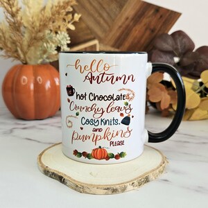 Puede incluir: Taza de cer&aacute;mica blanca con asa negra. La taza tiene un borde negro y presenta un dise&ntilde;o de oto&ntilde;o con el texto "hello autumn, hot chocolate, crunchy leaves, cosy knits, and pumpkins please".