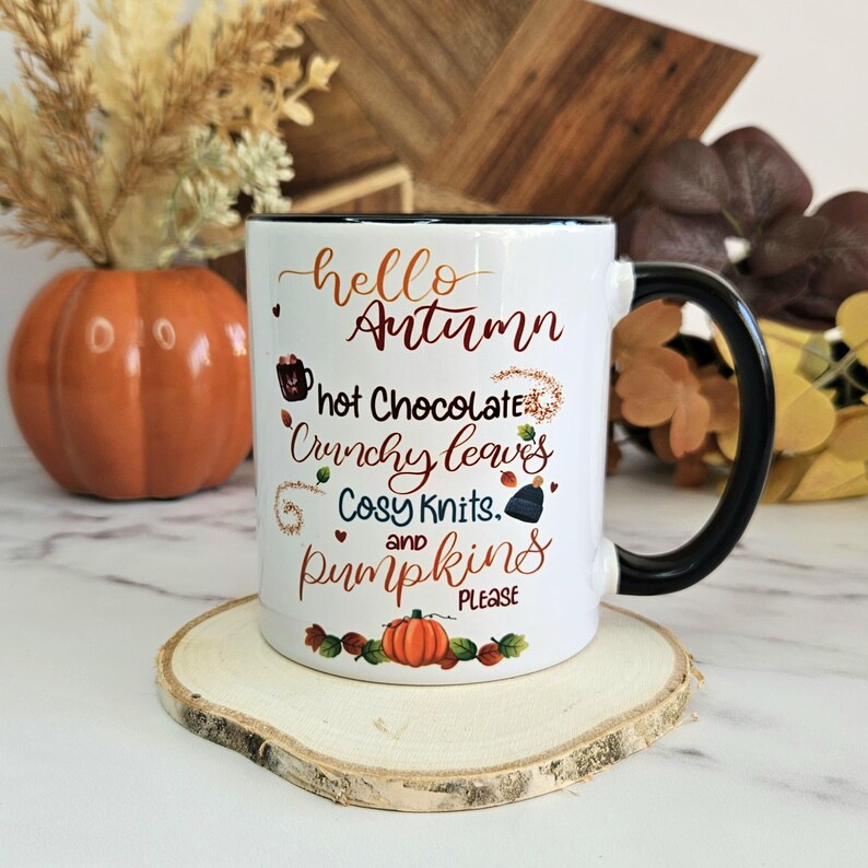 Puede incluir: Taza de cer&aacute;mica blanca con asa negra. La taza tiene un borde negro y presenta un dise&ntilde;o de oto&ntilde;o con el texto "hello autumn, hot chocolate, crunchy leaves, cosy knits, and pumpkins please".