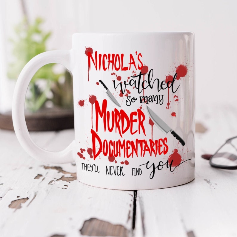 Op de afbeelding: Witte keramische koffiemok met een rode afbeelding waarop staat "Nicholas's watched so many murder documentaries they'll never find you". De afbeelding bevat twee messen en rode bloedspatten.