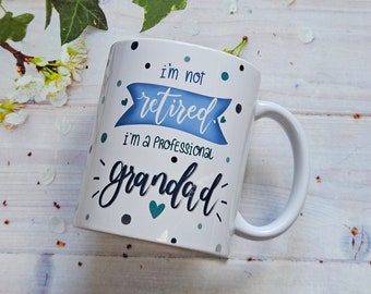 May include: White ceramic mug with the text "I'm not retired, I'm a professional grandad." The text is in a mix of blue and black fonts, with decorative dots and hearts. The mug has a curved handle.