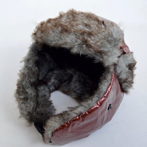 May include: A brown faux fur aviator hat with a shiny burgundy shell. The hat has a large fur brim and a small black button on the side.