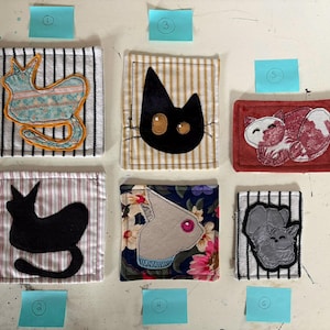 May include: A collection of handmade fabric coasters featuring various cat and animal designs. Each coaster has a unique design, including a black cat, a sleeping fox, and a squirrel. The coasters are square and have striped or floral backgrounds.