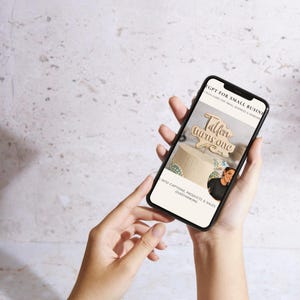 May include: A smartphone screen showing the text "GPT for Small Business" and "Taller turns one." The image on the screen is of a wooden cake topper. The phone is held in two hands against a neutral background.