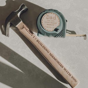 May include: A metal hammer with a wooden handle engraved with the text DADDY I LOVE BUILDING MEMORIES WITH YOU and a teal tape measure featuring a circular label that reads Dad You are loved beyond measure.