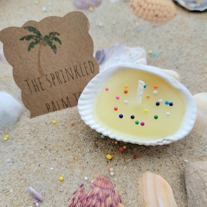 May include: A seashell candle with a yellow wax interior and colorful sprinkles. A white wick is centered in the candle. The candle sits on sand with seashells and a brown tag that reads "The Sprinkled Palm Tree" with a palm tree graphic.