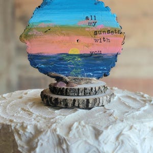 May include: A cake topper featuring a painted sunset scene on a wood slice. The sunset has blue, pink, and yellow hues, with the words "all my sunsets with you." Two small wood slices and seashells are at the base.