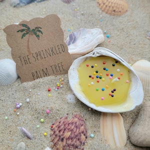 May include: A yellow candle with colorful sprinkles sits inside a seashell on a sandy surface. A brown tag with a palm tree illustration and the text "THE SPRINKLED PALM TREE" is next to the candle. Seashells and colorful sprinkles surround the candle.