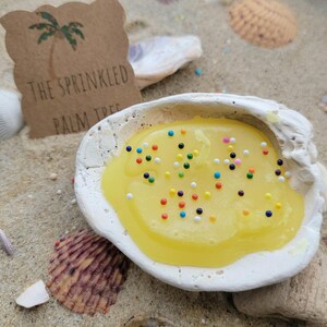 May include: A white seashell filled with yellow liquid and colorful sprinkles. A brown tag with a palm tree illustration and the text "THE SPRINKLED PALM TREE" is in the background. The scene is set on sand with seashells.