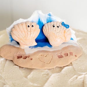 May include: A whimsical cake topper featuring two smiling seashell figures with googly eyes, set against a blue and white wave design. A sandy base with a heart and the word "Mrs." completes the beachy theme.