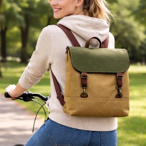 May include: A canvas backpack with a green flap and brown straps. The bag has a tan body and brown leather-like straps with metal hardware. The backpack is being worn by a person riding a bicycle.