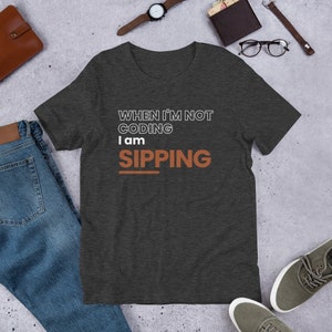 Op de afbeelding: Donkergrijs t-shirt met de witte en oranje tekst "WHEN I'M NOT CODING I am SIPPING". Het shirt wordt getoond op een getextureerd oppervlak met jeans, schoenen en accessoires.