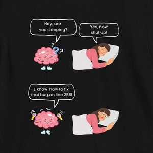 Op de afbeelding: Zwart T-shirt met een humoristische afbeelding. De afbeelding toont een roze brein dat vraagt: "Hey, are you sleeping?" en een persoon in bed die antwoordt: "Yes, now shut up!" Daaronder zegt het brein: "I know how to fix that bug on line 255!"