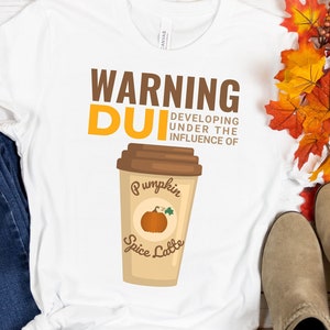 以下が含まれることがあります： 「WARNING DUI DEVELOPING UNDER THE INFLUENCE OF Pumpkin Spice Latte」の文字が入った白いTシャツ。青いジーンズ、コーヒーカップ、カボチャや葉など、秋をテーマにした装飾が施されています。