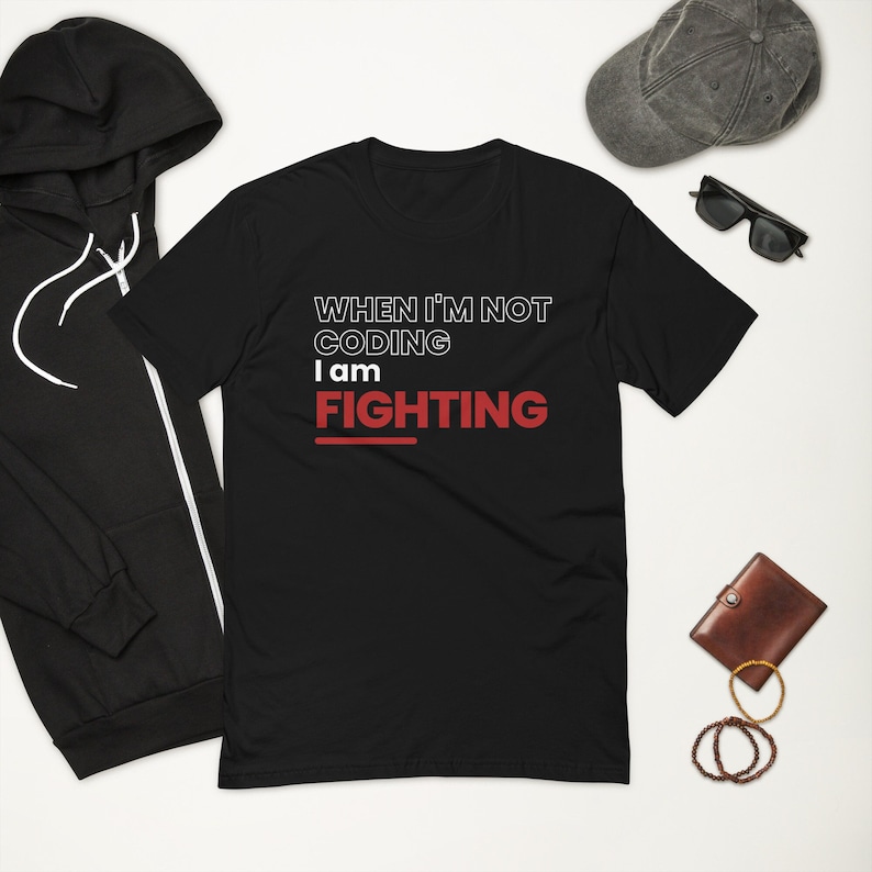 K&ouml;nnte beinhalten: Schwarzes T-Shirt mit dem Text "WHEN I'M NOT CODING I AM FIGHTING" in Wei&szlig; und Rot. Das Bild enth&auml;lt auch einen schwarzen Kapuzenpullover, eine graue Kappe, Sonnenbrillen, eine braune Geldb&ouml;rse und Armb&auml;nder.