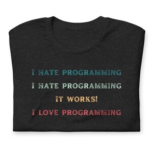 Puede incluir: Camiseta gris oscuro con el texto "I HATE PROGRAMMING" en turquesa, "I HATE PROGRAMMING" en turquesa claro, "IT WORKS!" en dorado y "I LOVE PROGRAMMING" en rojo. La camiseta está doblada.