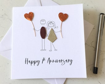May include: A white card with a hand-drawn illustration of a couple holding red heart balloons. The text "Happy 7th Anniversary" is written below the illustration.