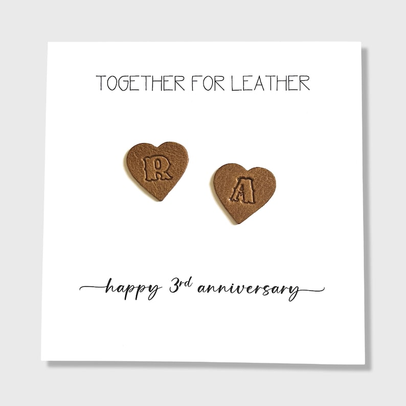 May include: A white card with the text "TOGETHER FOR LEATHER" and "happy 3rd anniversary". Two brown heart-shaped leather pieces are on the card, one with the letter "R" and the other with the letter "A".